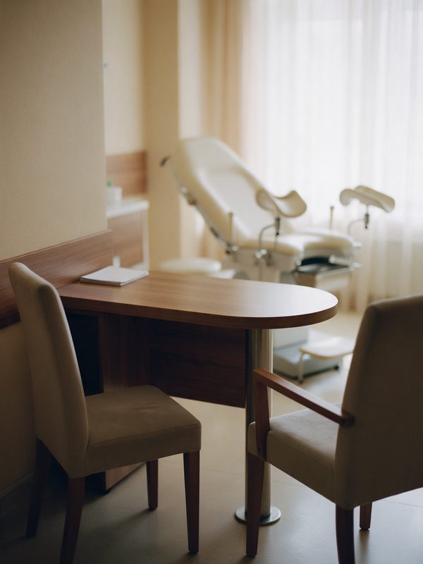 Consultório de ginecologia com mesa, duas cadeiras e a cadeira de exame ao fundo.