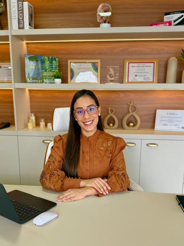 Dra. Joyce Menezes sentada à mesa do consultório, sorrindo para a câmera.
